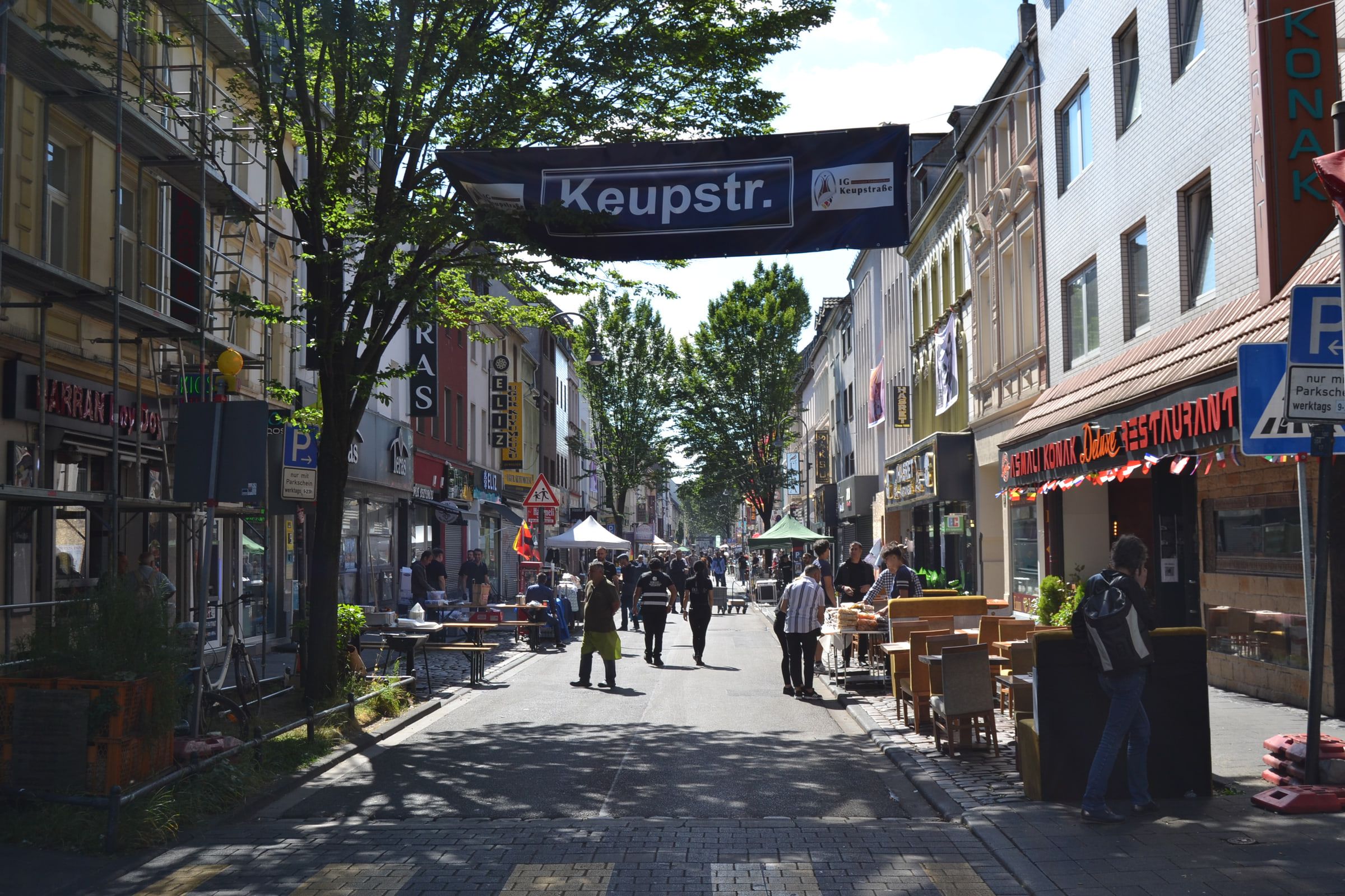 Die Keupstraße als Festmeile: ein Banner „Keupstr.“ der IG Keupstraße hängt zwischen den Häusern, darunter Tische, Zelte und Menschen auf der gesperrten Straße, links ein eingerüstetes Haus.