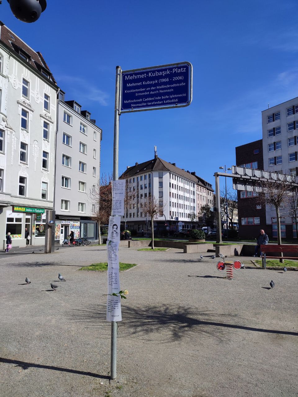 Ein blaues Straßenschild „Mehmet-Kubaşık-Platz“ mit Erläuterung auf Deutsch und Türkisch: Mehmet Kubaşık (1966–2006), Kioskbetreiber an der Mallinckrodtstraße, ermordet durch Neonazis. Am Mast hängen beschriebene Blätter und eine Rose; dahinter ein Platz mit Spielgerät und Wohnhäusern.