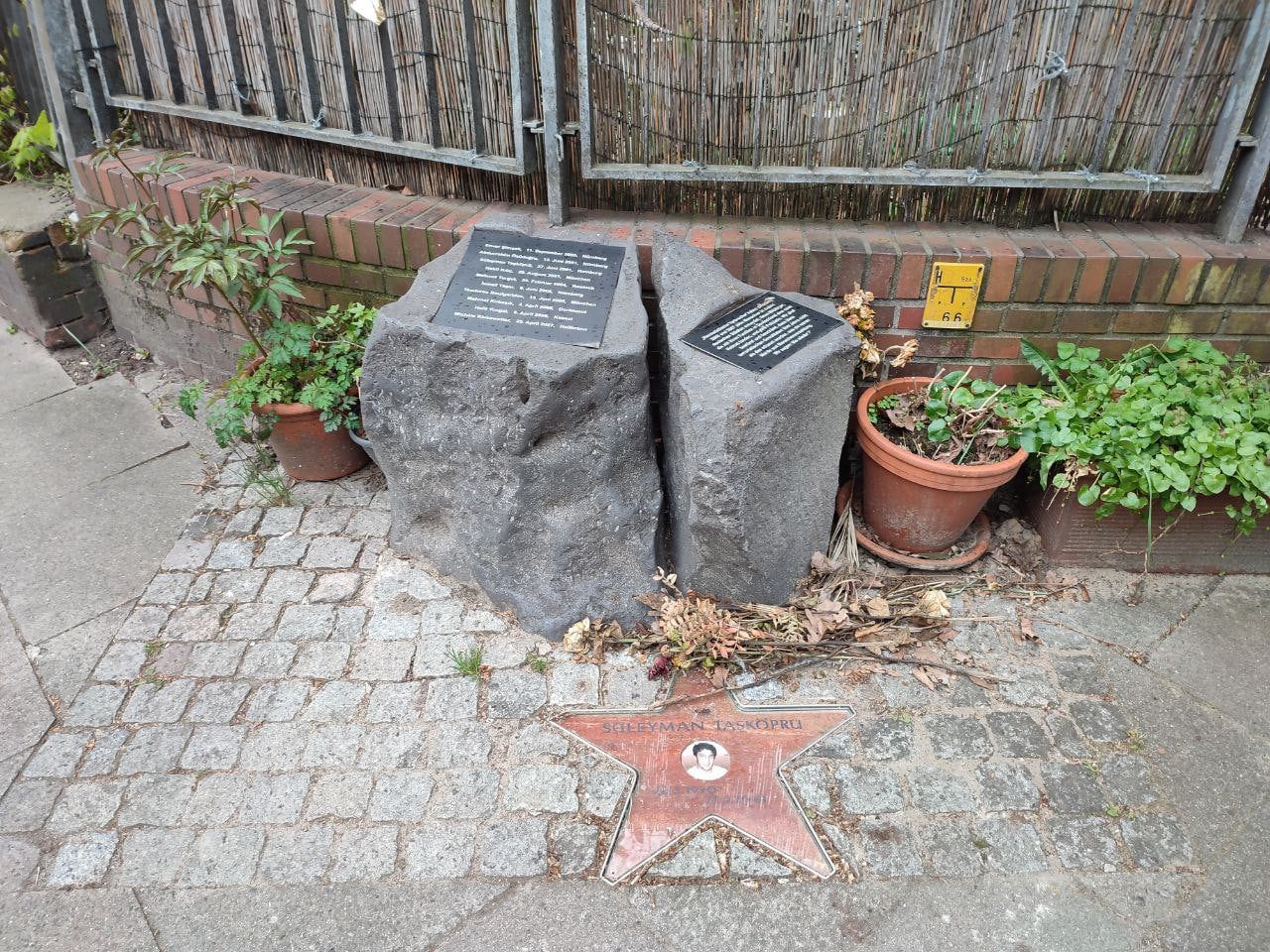 Zwei Gedenksteine an einer niedrigen Backsteinmauer, der linke mit einer Tafel, die die Namen der zehn Mordopfer des NSU mit Datum und Ort aufzählt; davor im Pflaster ein Stern mit dem Namen Süleyman Taşköprü und seinem Porträt, daneben Blumentöpfe.